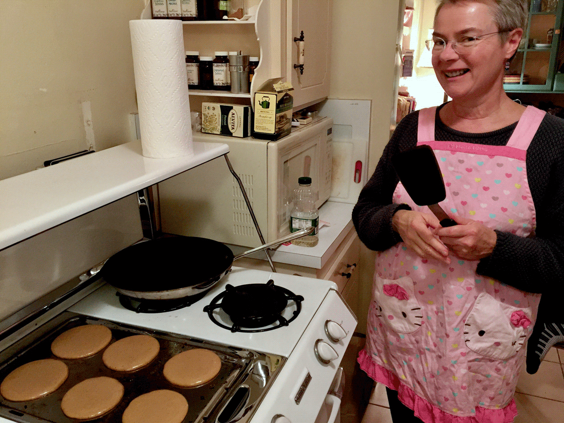 Linda hoding spatula in Hello Kitty apron with six pancakes cooking on the griddle